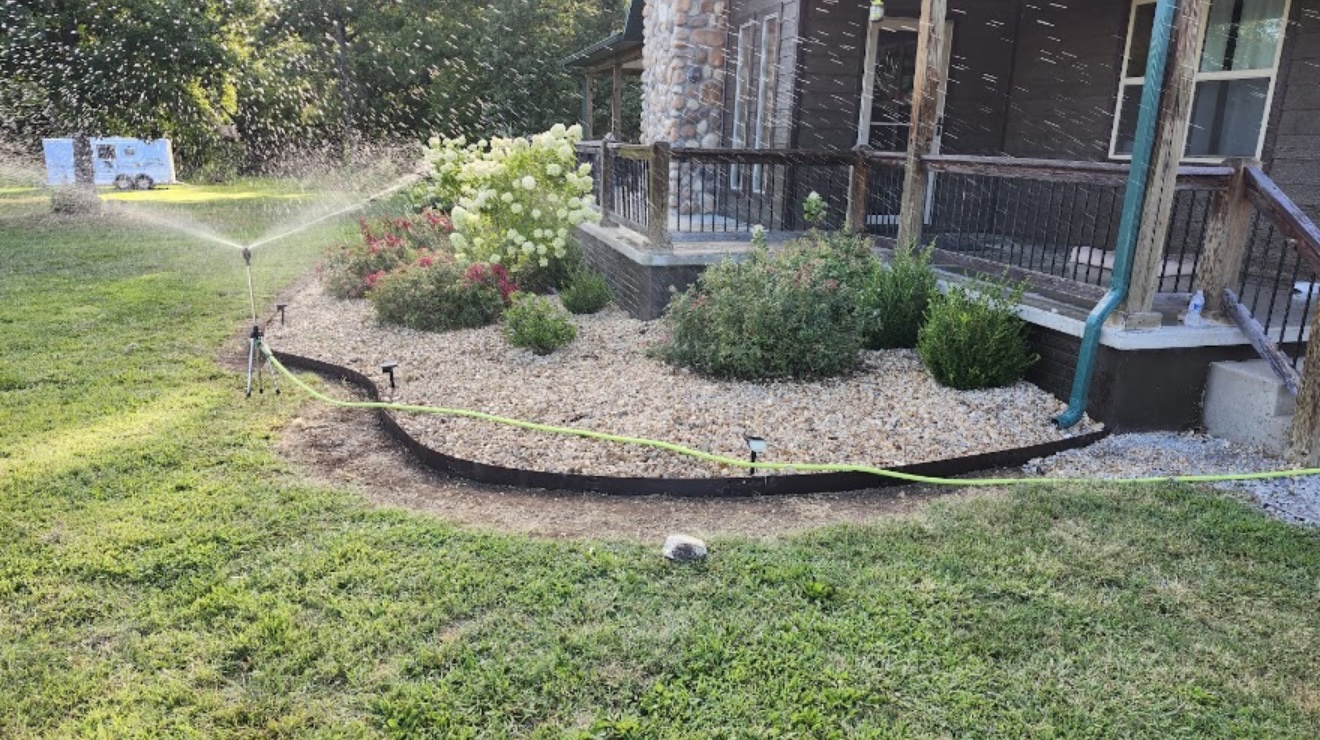 New landscape bed with gravel, shrubs, and flowers along a house porch while a sprinkler waters the lawn
